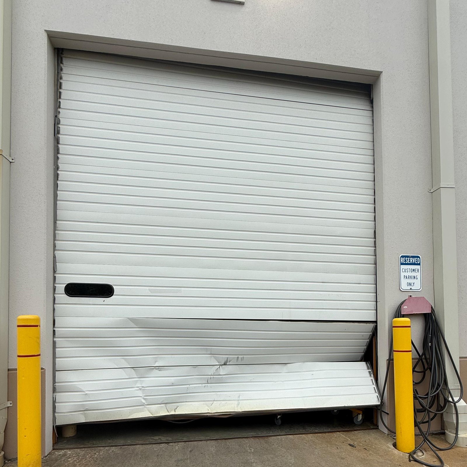 Commercial overhead door with its bottom sections dented and bent by an impact, between two yellow safety posts – Joy Garage Services job