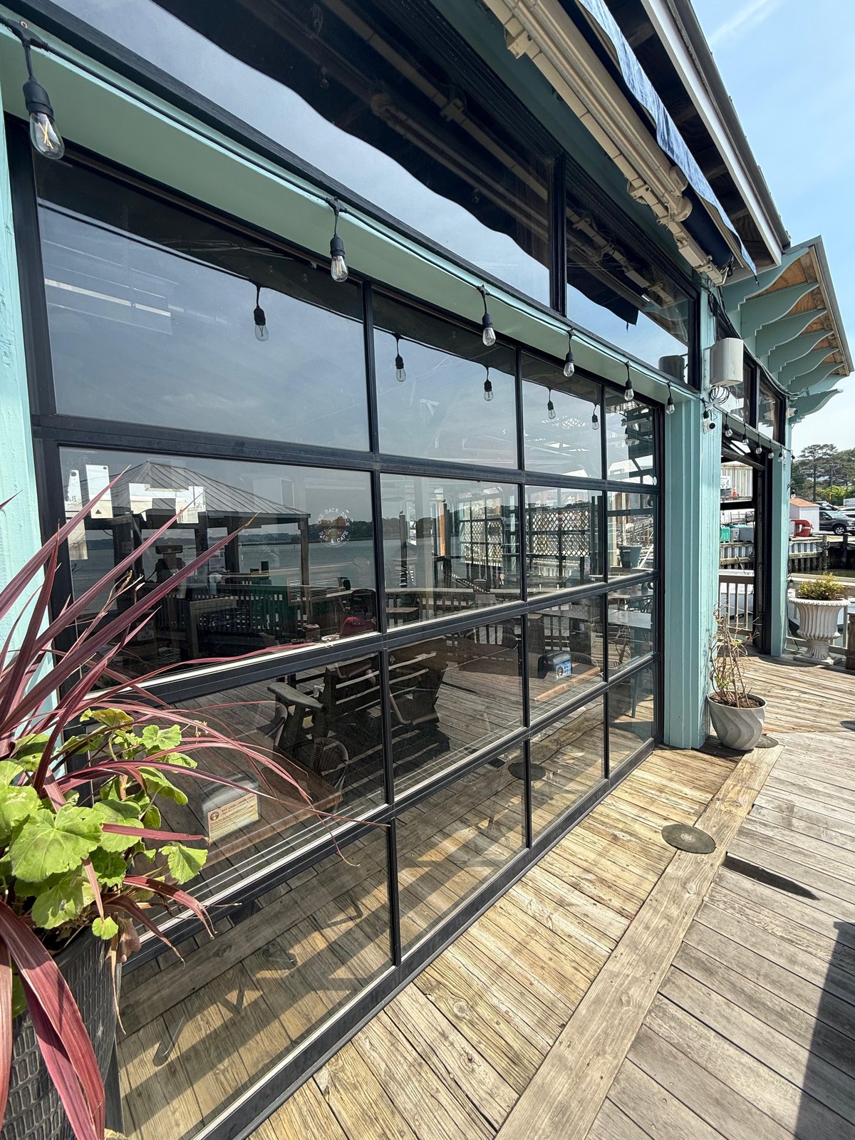 Full-view glass garage door with a black aluminum frame, next to a wooden deck with plants