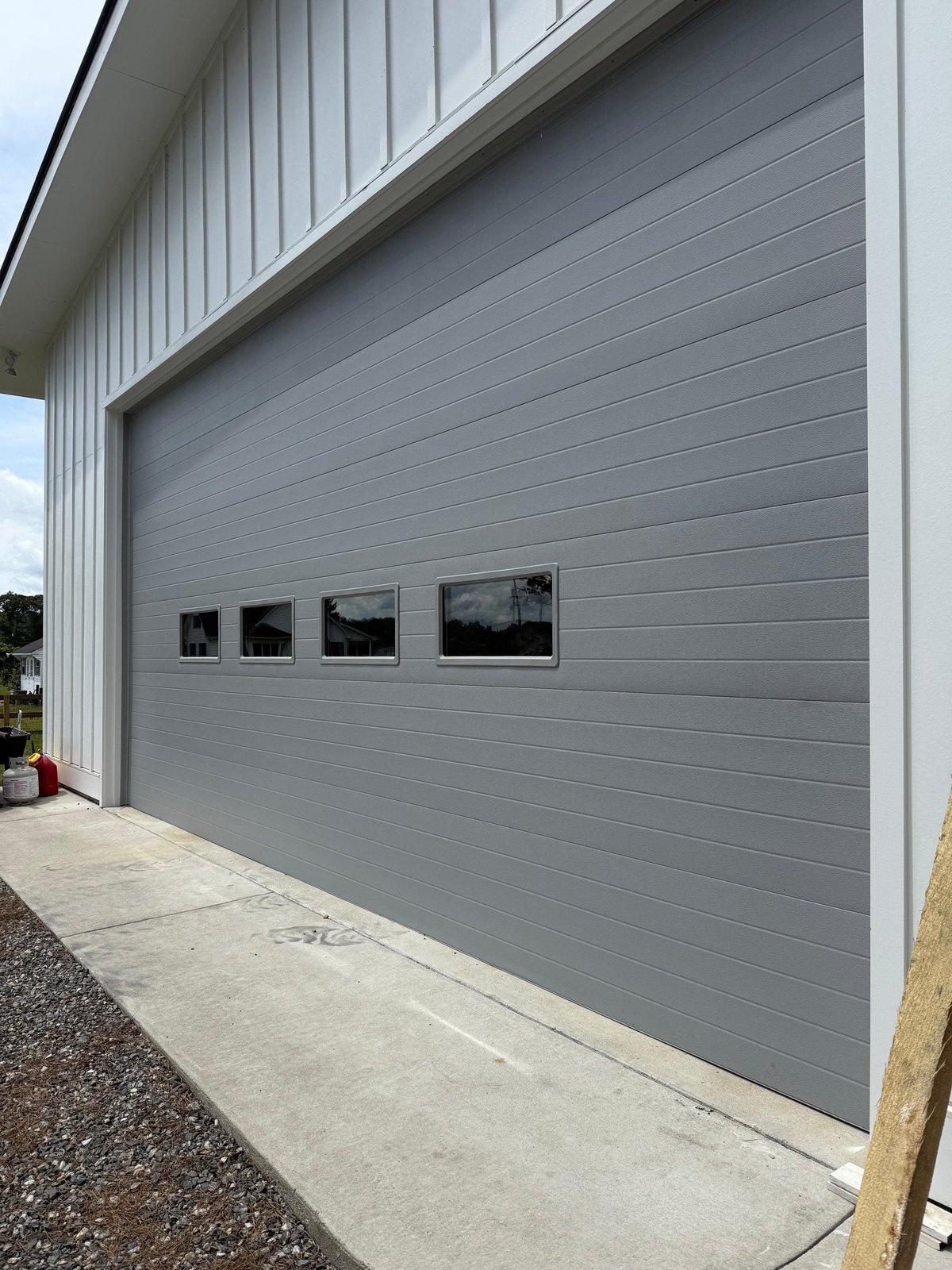 Large gray flush garage door with a row of small windows on a white board-and-batten building