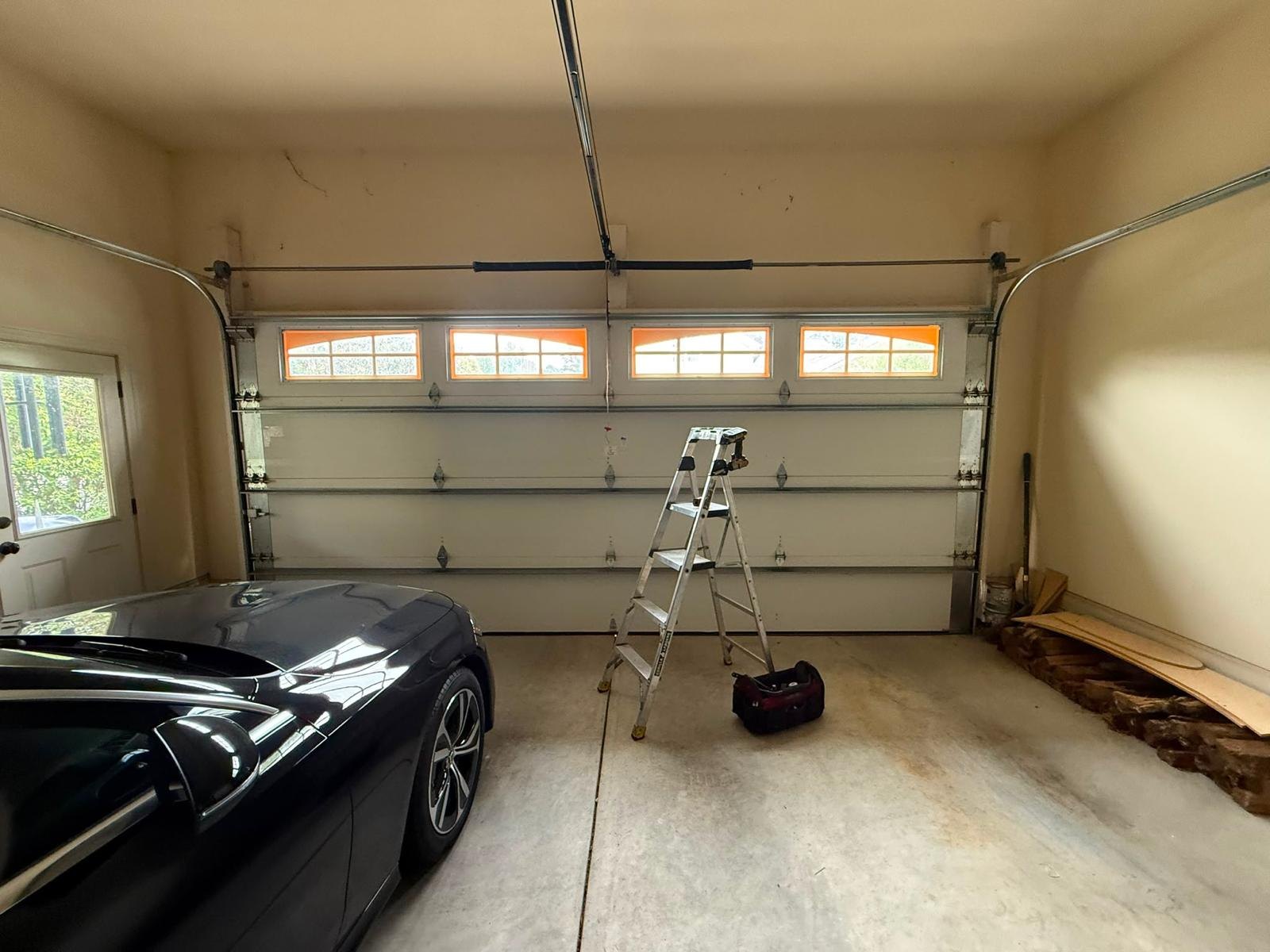 Inside a garage: a white door with a row of windows, the torsion spring on the bar above it, a stepladder and a dark car