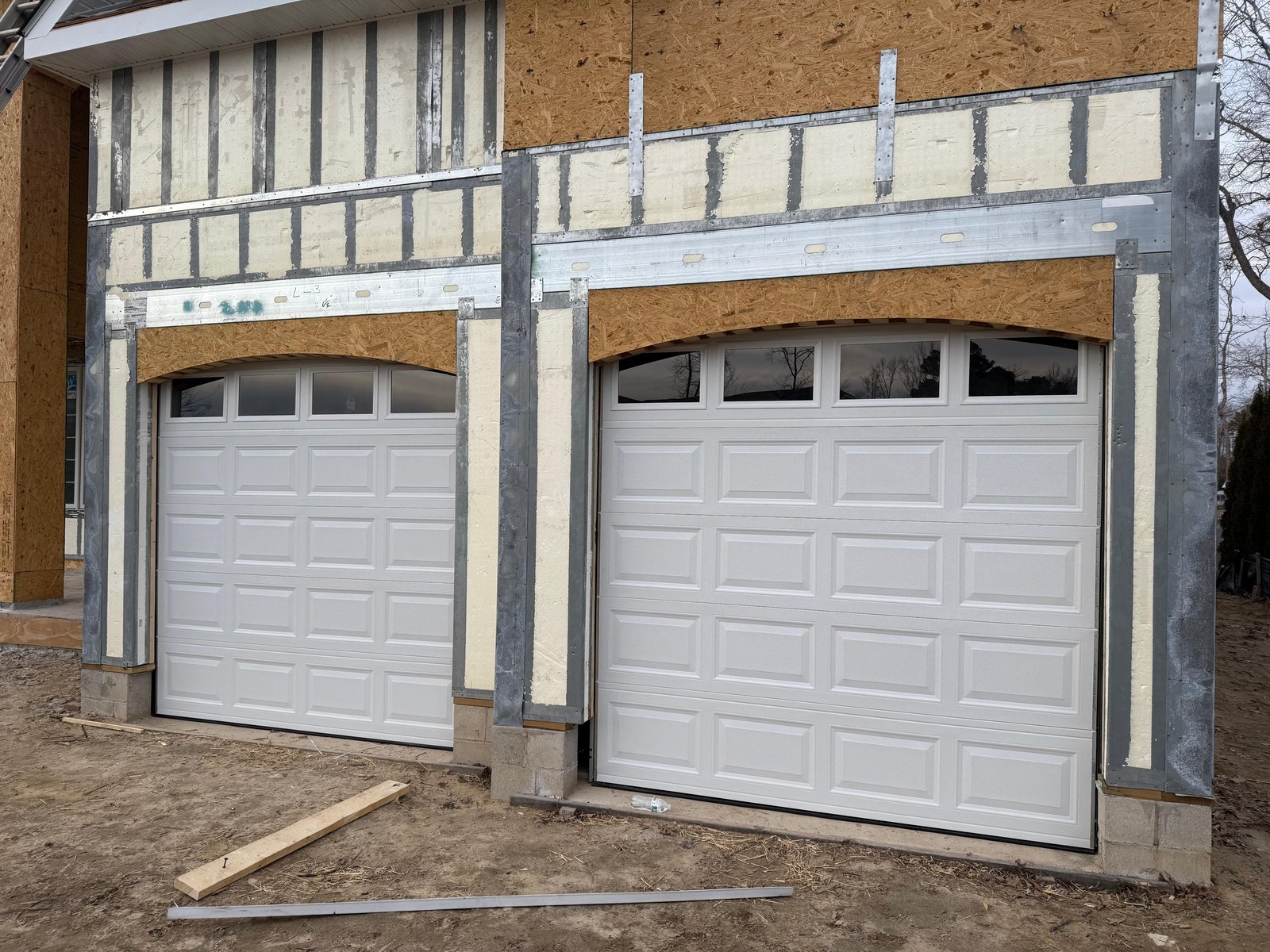 Two new white raised-panel garage doors with a row of windows, in the openings of a home under construction